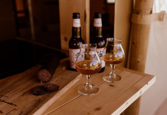 Twee glazen en bierflesjes met worst op houten tafel in safaritent, ontspannen sfeer en daglicht.