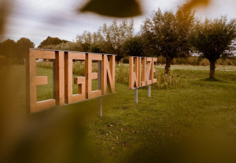 Insegna in legno 'EIGEN WIJZE' su un prato al parco vacanze Eigen Wijze nei Paesi Bassi, con alberi sullo sfondo.