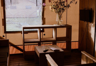 Cozy safari tent interior with wooden furniture, dried flowers in a vase, and muted light from a window.