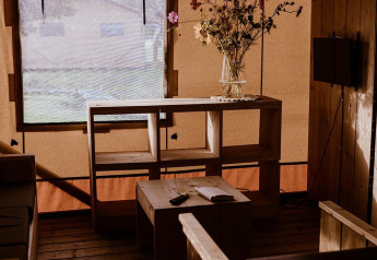 Cozy safari tent interior with wooden furniture, dried flowers in a vase, and muted light from a window.
