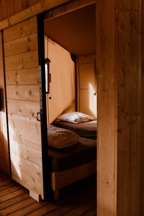Cozy safari tent bedroom with wooden walls, a comfortable bed, and soft sunlight streaming through.