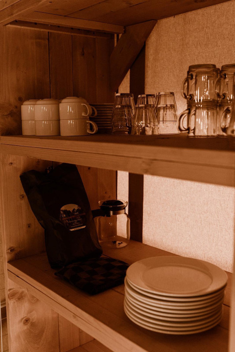 Wooden shelf in a safari tent kitchen with stacked plates, mugs, glasses, and a coffee bag in warm light.