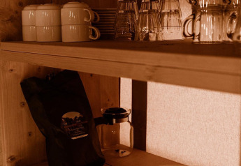 Wooden shelf in a safari tent kitchen with stacked plates, mugs, glasses, and a coffee bag in warm light.