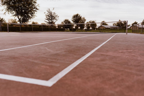 Un court de tennis extérieur vide avec surface rougeâtre et lignes blanches, entouré d’arbres et de clôtures.