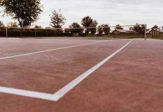 An empty outdoor tennis court with a reddish surface and white lines, surrounded by trees and fencing.