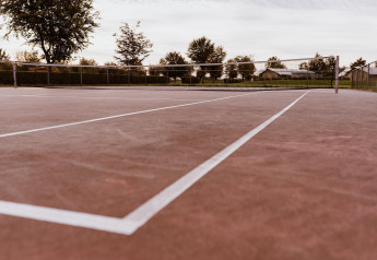 An empty outdoor tennis court with a reddish surface and white lines, surrounded by trees and fencing.