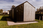 Modernes Glamping-Haus mit Holzverkleidung, umgeben von grünem Rasen und Bäumen bei Sonnenschein.
