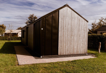 Modernes Glamping-Haus mit Holzverkleidung, umgeben von grünem Rasen und Bäumen bei Sonnenschein.