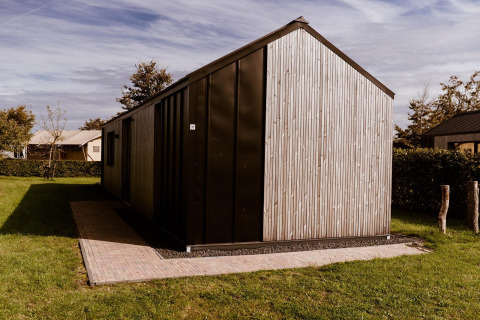 Moderne glampinghut met houten en donkere panelen, omgeven door gras en bomen onder een heldere hemel.