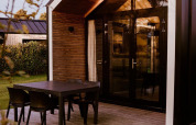 Moderne glamping cabin met grote glazen deuren, houten terras en zwarte tafel met stoelen buiten.