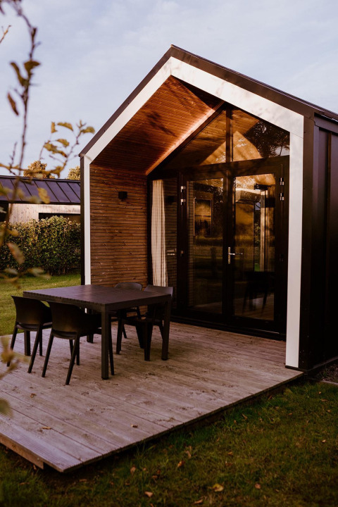 Cabane de glamping moderne avec baies vitrées, terrasse en bois et table noire dans un cadre naturel.