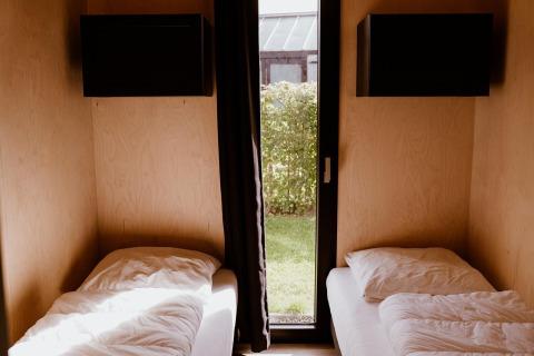 Twee eenpersoonsbedden in een houten glamping accommodatie met groot raam en zicht op een groene tuin.