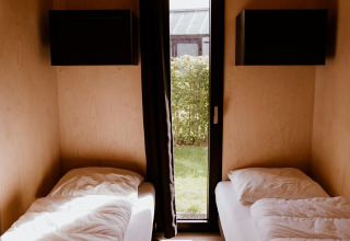 Dos camas individuales en una luminosa cabaña glamping de madera con ventanas que miran a un jardín verde.