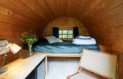 Acogedora cabaña de madera con techo curvo, cama, flores en jarrón y luz natural por la ventana.