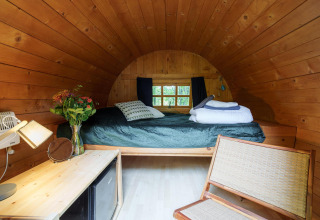 Cozy wooden cabin with curved ceiling, bed, flowers in vase, natural light, and minimalist decor.
