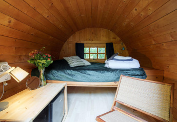 Acogedora cabaña de madera con techo curvo, cama, flores en jarrón y luz natural por la ventana.