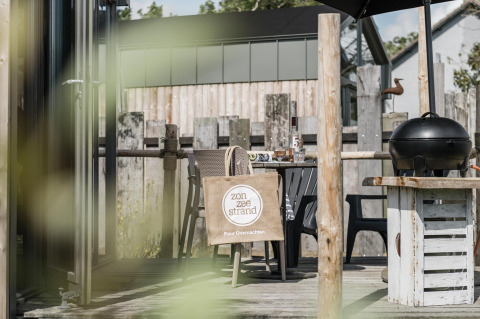 Terrasse am Rescue cottage im Duynpark Het Zwanenwater, Niederlande, mit Tisch, Grill und Einkaufstasche.