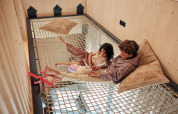 Dos niños se relajan en una red suspendida con cojines y tablet en Unbrick One | Sauna en Brinckerduyn, Países Bajos.