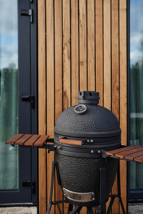 Parrilla de cerámica negra con termómetro frente a paneles de madera en una moderna cabaña con puerta de vidrio.