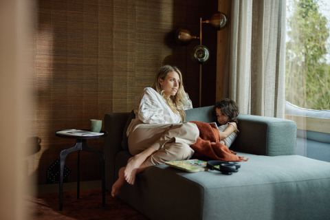 A woman and child relax together on a sofa by the window in a cozy lodge with a nature view outside.