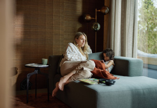 A woman and child relax together on a sofa by the window in a cozy lodge with a nature view outside.