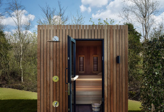 Buitensauna bij een lodge met houten gevel, deur open en zitbank zichtbaar, omgeven door groene natuur.