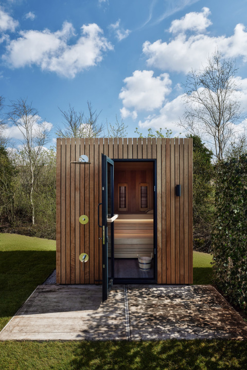 Sauna de madera exterior junto a una cabaña, puerta abierta con banco visible, rodeada de naturaleza verde.