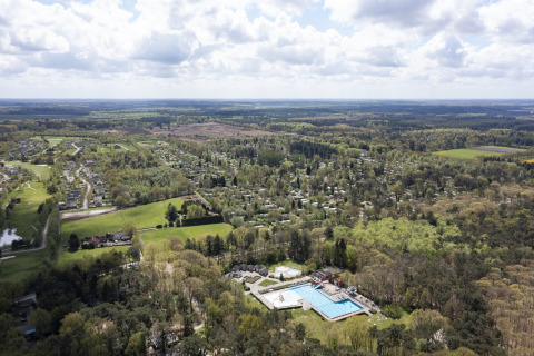 Vista aérea de un complejo vacacional con piscina, casas dispersas y bosque bajo un cielo parcialmente nublado.