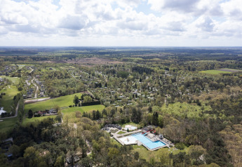 Vista aérea de un complejo vacacional con piscina, casas dispersas y bosque bajo un cielo parcialmente nublado.