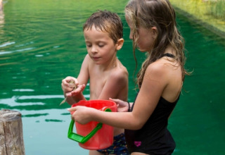 Dos niños junto a un lago en Eco domaine Le Camp, Occitania, Francia, sostienen un cubo y una rana pequeña.