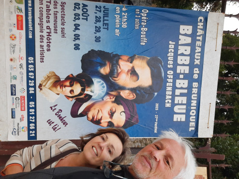 Dos personas sonrientes frente a un cartel de Barbe-Bleue de Offenbach en Châteaux de Bruniquel, Occitanie.