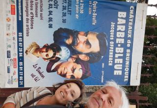 Deux personnes souriantes devant une affiche de Barbe-Bleue d'Offenbach aux Châteaux de Bruniquel, Occitanie.