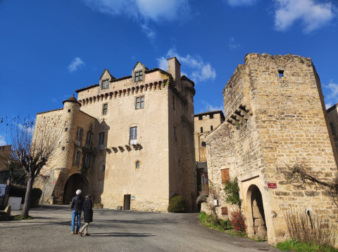 Zwei Personen spazieren vor einer historischen Steinburg unter blauem Himmel in der Nähe von Varen, Occitanie.