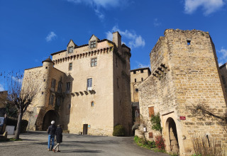 Zwei Personen spazieren vor einer historischen Steinburg unter blauem Himmel in der Nähe von Varen, Occitanie.