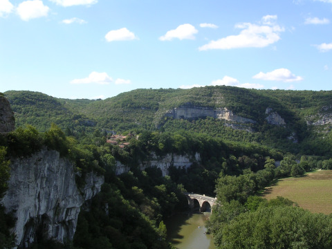 Paisaje en Eco domaine Le Camp, Occitanie, Francia, con colinas boscosas, río, puente de piedra y cielo azul.