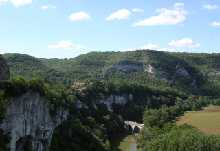 Paisaje en Eco domaine Le Camp, Occitanie, Francia, con colinas boscosas, río, puente de piedra y cielo azul.