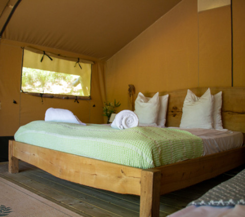 Vista interior de una tienda safari con cama de madera, mantas verdes y toallas blancas enrolladas.