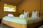 Vista interior de una tienda safari con cama de madera, mantas verdes y toallas blancas enrolladas.