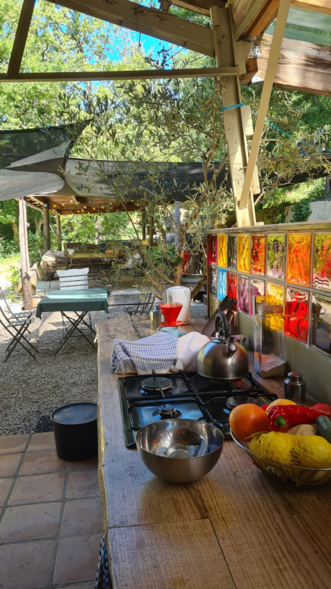 Outdoor-Küche am Safarizelt mit buntem Fenster, Obst und Sitzbereich unter Bäumen im Freien.