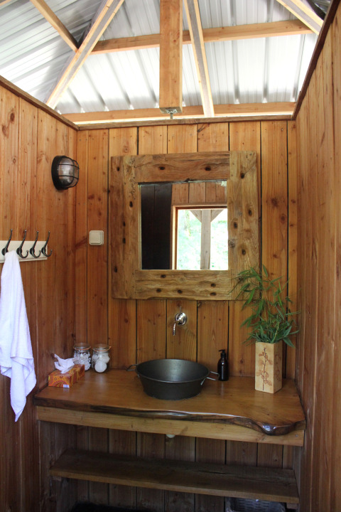 Safari-Zelt-Badezimmer mit Holzverkleidung, rustikalem Spiegel, Metallschüssel und Pflanze.