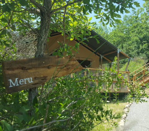 A safari tent named Lodgetent Meru is partially hidden behind green foliage, with a wooden sign in front.