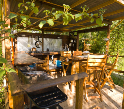 Cocina exterior rústica y zona de comedor de madera rodeada de vegetación, en Le Camp, Francia.