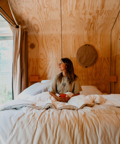Vrouw zit in bed met een tas, omgeven door licht hout in Les Cabanes d'Ostende, West-Vlaanderen.
