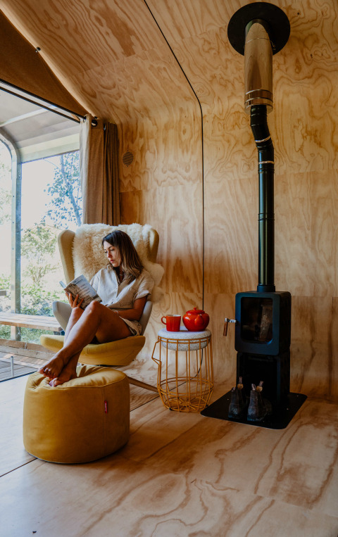 Femme lisant dans une cabane en bois cosy avec poêle et mobilier jaune, Les Cabanes d'Ostende, Belgique.