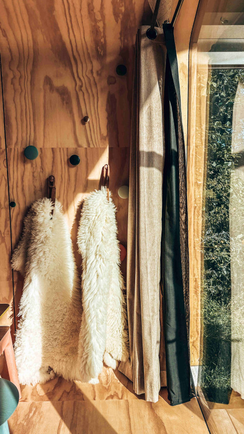 Sunlight streams on wooden wall, curtains, and two white sheepskins at Les Cabanes d'Ostende, West Flanders.