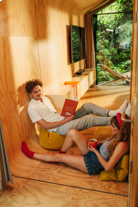 Dos personas se relajan en una cabaña moderna de madera con ventanales y vistas a la naturaleza verde.
