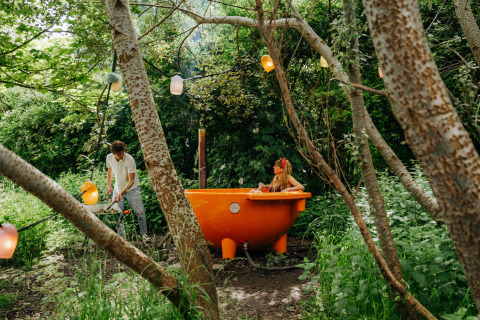 Un hombre calienta una bañera naranja al aire libre mientras una mujer se relaja rodeada de bosque.