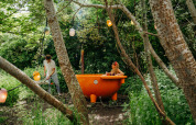 Un hombre calienta una bañera naranja al aire libre mientras una mujer se relaja rodeada de bosque.
