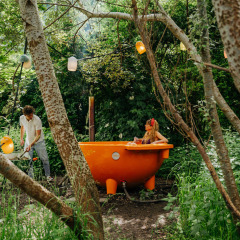 Un hombre calienta una bañera naranja al aire libre mientras una mujer se relaja rodeada de bosque.