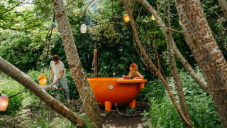 Un hombre calienta una bañera naranja al aire libre mientras una mujer se relaja rodeada de bosque.